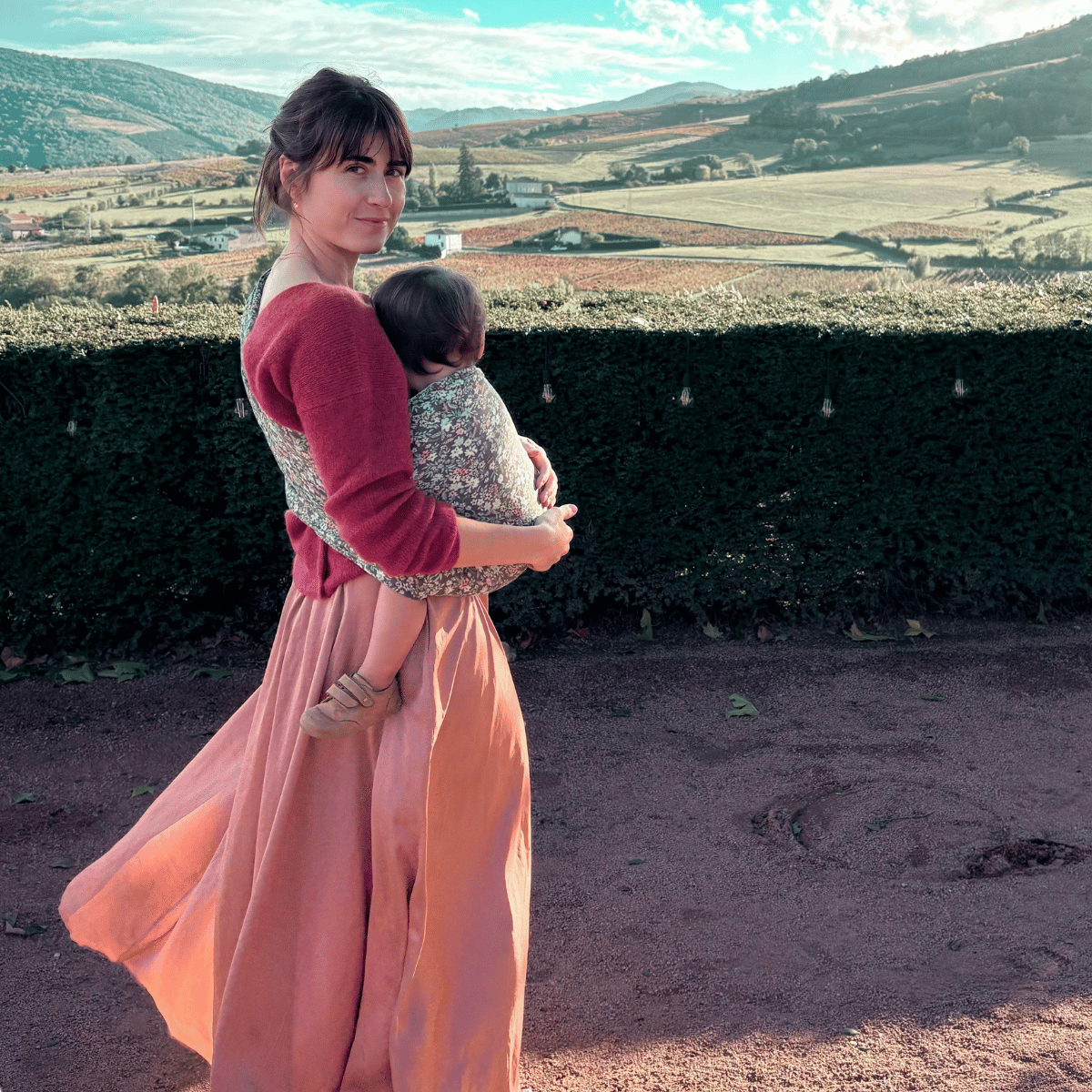 A light skinned brown haired straight sized woman stands outside and looks over her shoulder at the camera while she wear her light skinned brown haired baby in a hip carry in a ring sling. She stands in front of bucolic landscape.