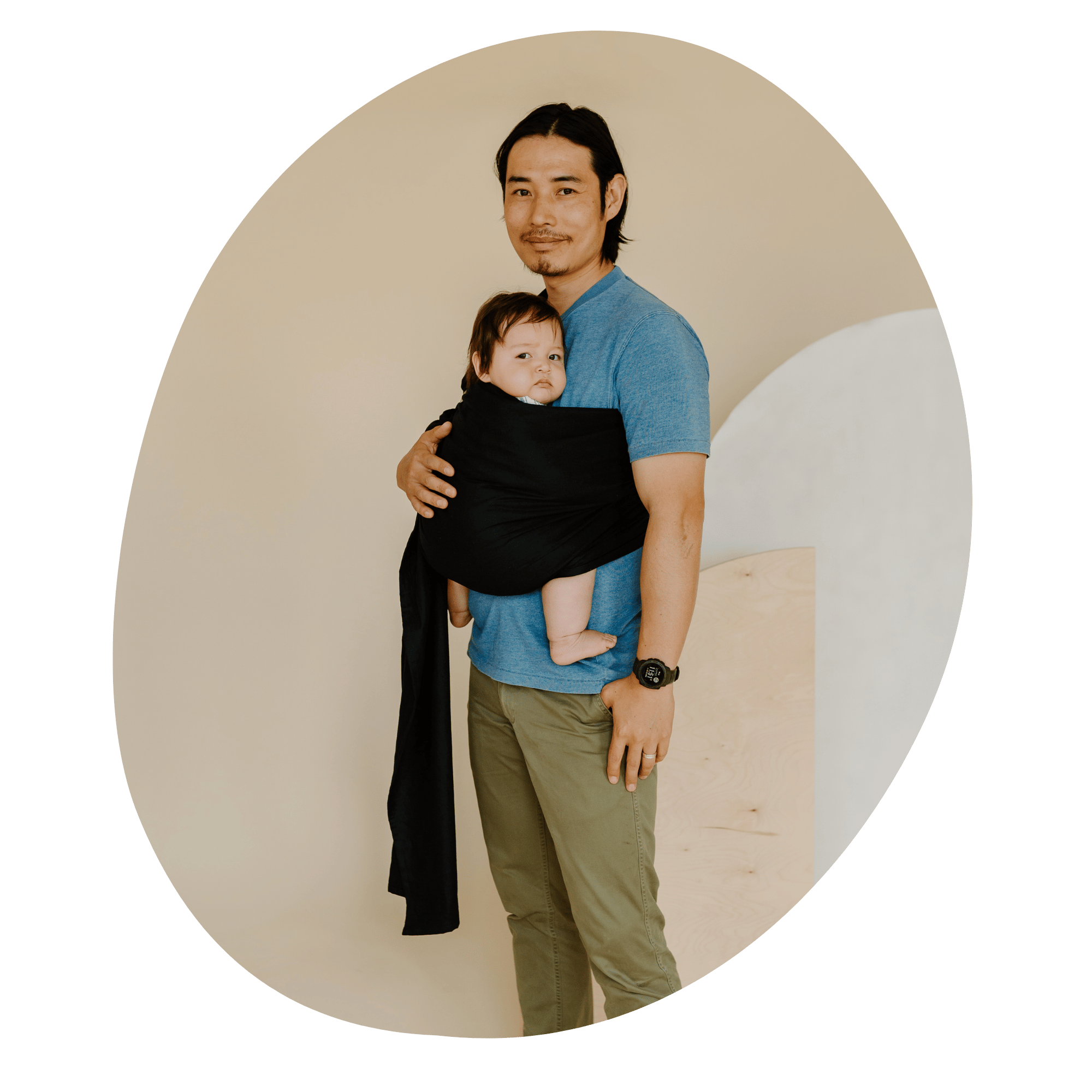 A BIPOC straight size man smiles at the camera while wearing his baby on his front in a ring sling.
