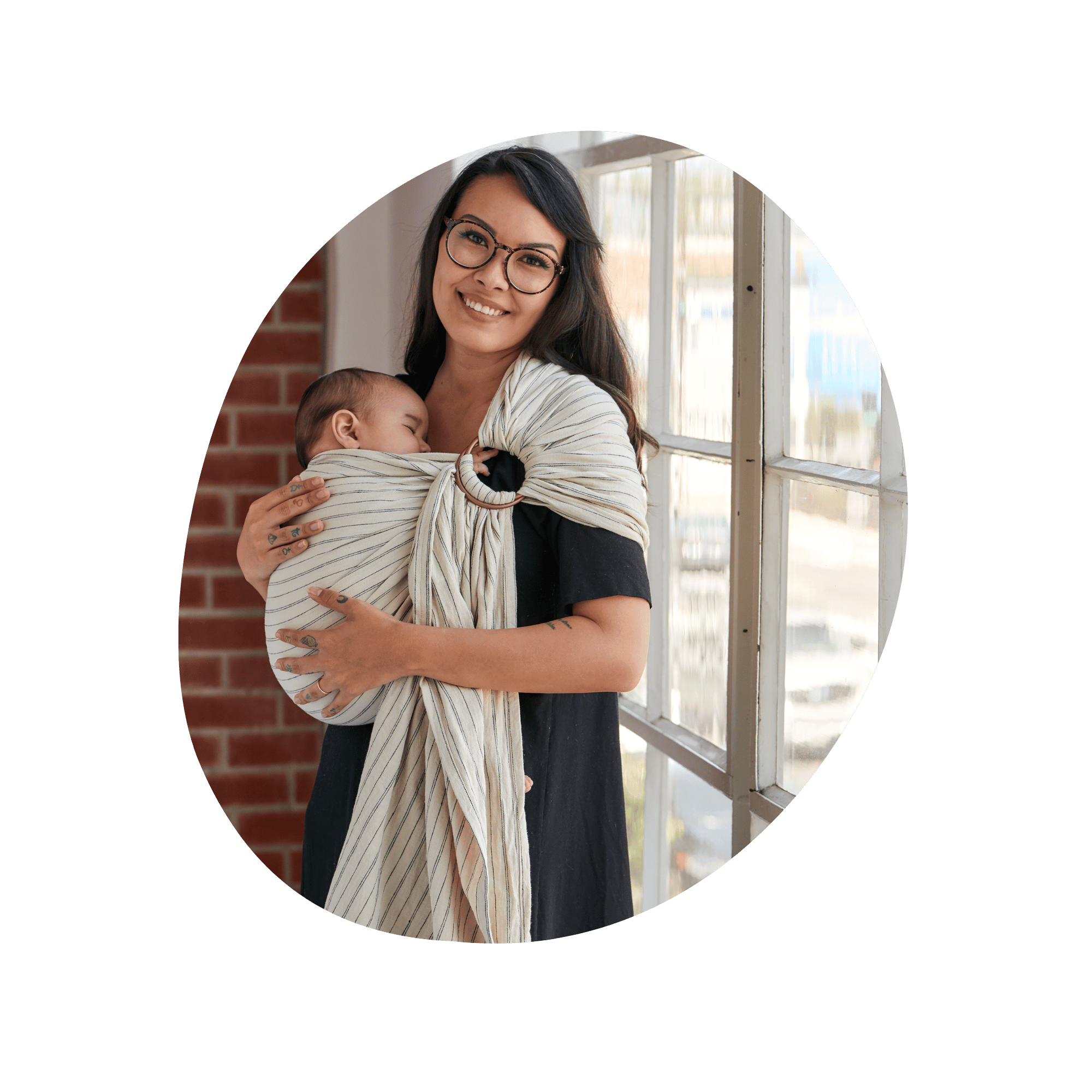 A woman smiles at the camera as she wears a sleeping baby in a ring sling