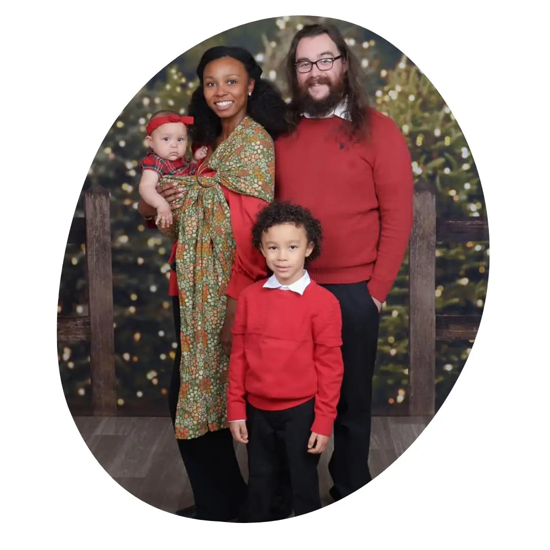a family of 4 in red sweaters stands in front of lights. the youngest is worn in a ring sling