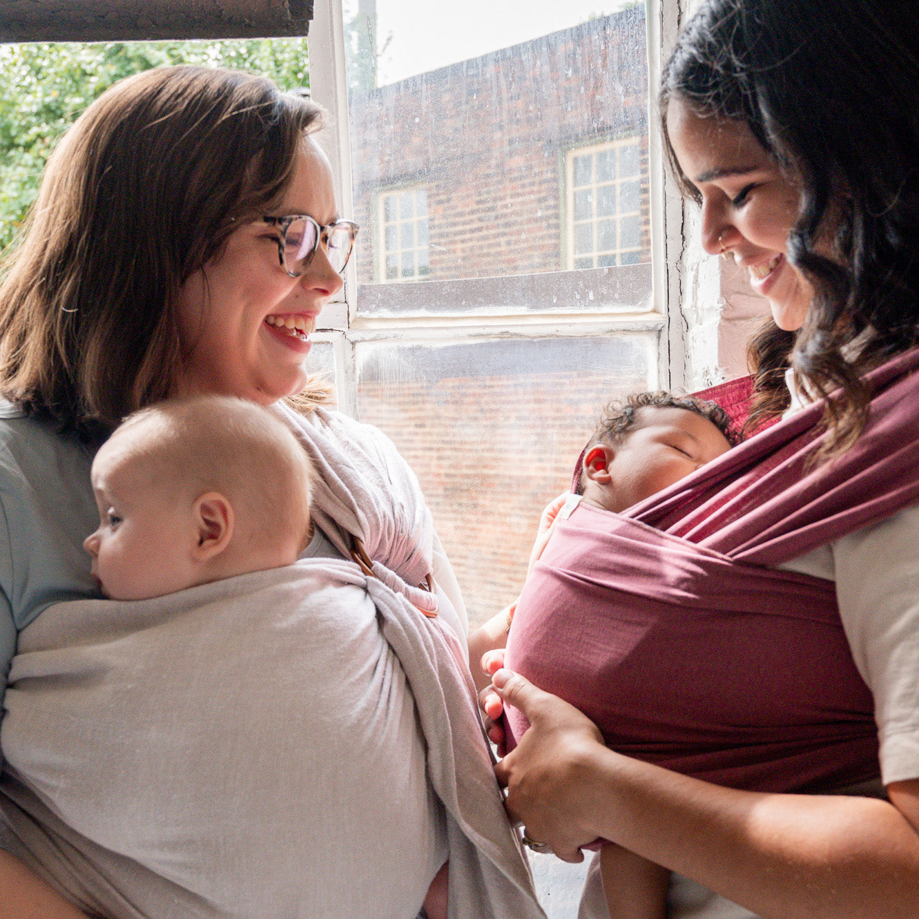 White mother wearing her baby in a gray ring sling talking to another mother wearing her newborn in a rose-colored baby wrap.