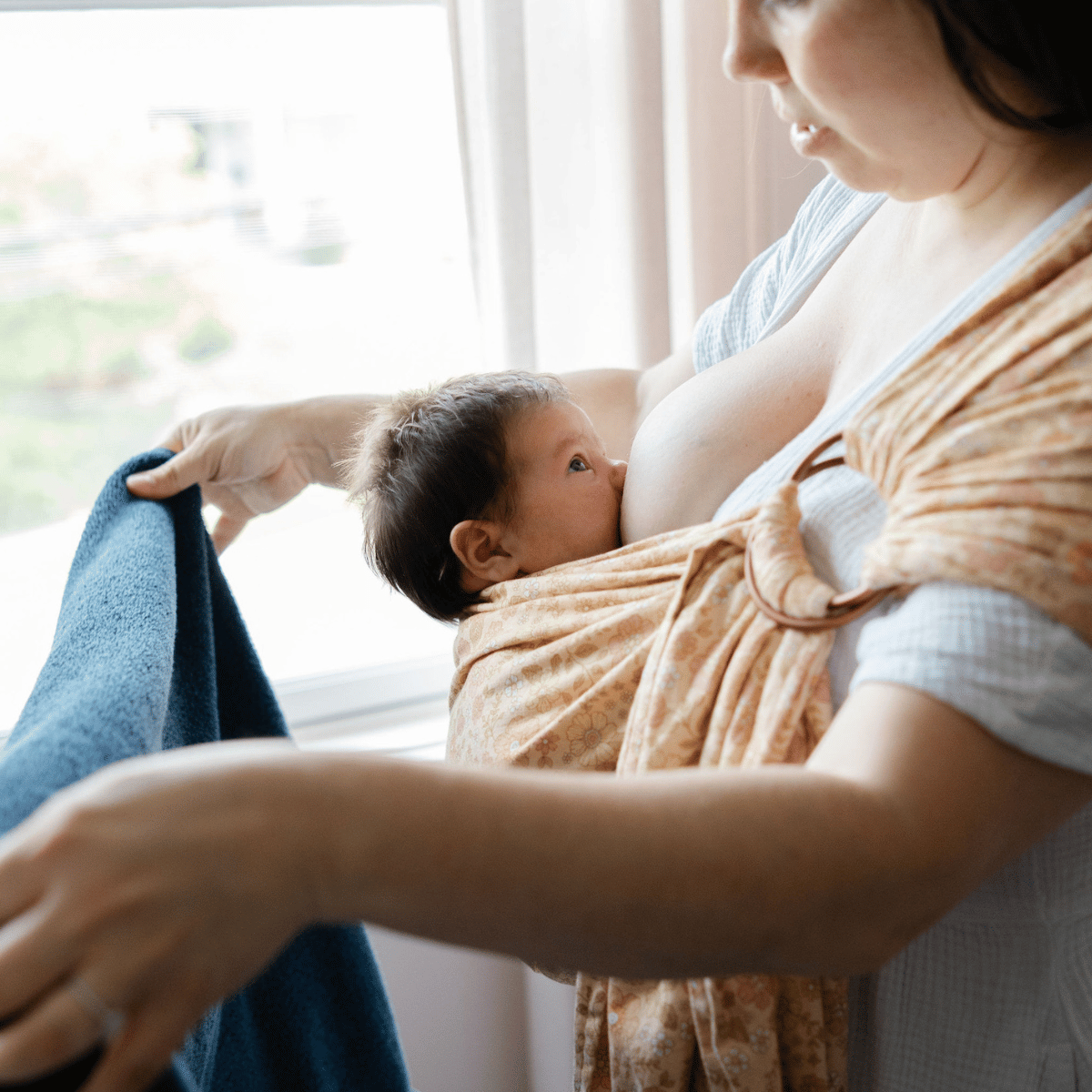 A mid size white woman wears a newborn in a hope&plum ring sling while nursing and folding laundry