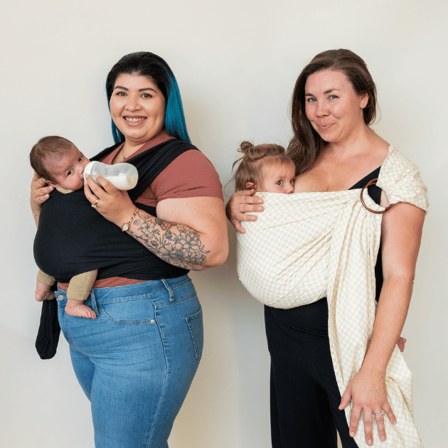 Two women stand in nearly profile. The first wears her baby in a stretchy wrap and bottle feeds. The second wears her baby in a ring sling and nurses.