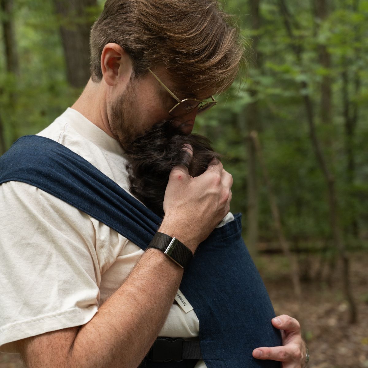 A straight size white man stands in profile and kisses their infant on the top of the head while wearing them in a hope&plum Caspian Sprout Newborn Carrier