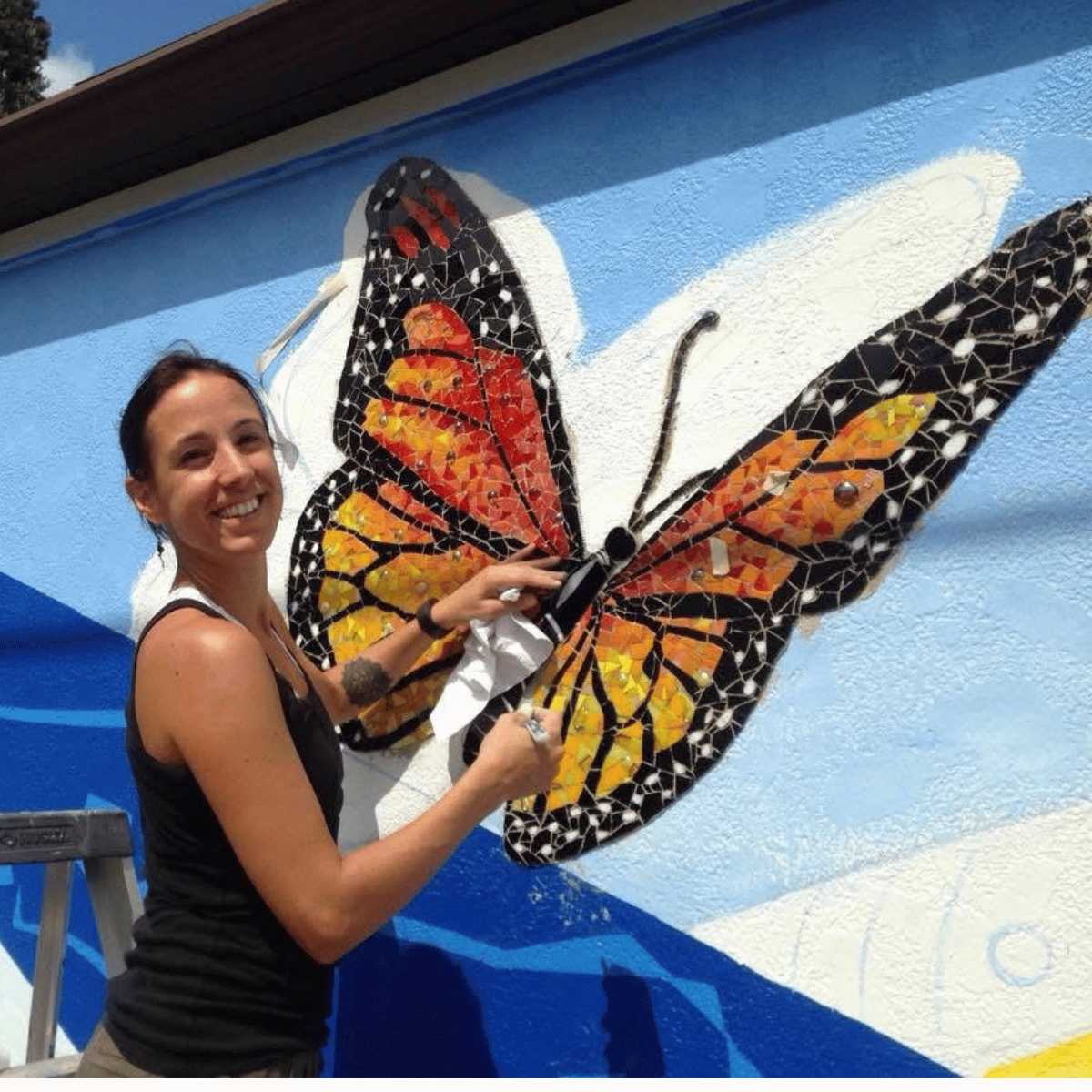 A straight size BIPOC woman turns to smile at the camera while working on a butterfly mosaic on the side of a building
