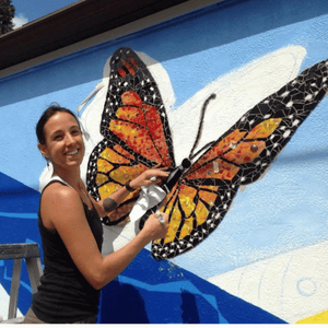 A straight size BIPOC woman turns to smile at the camera while working on a butterfly mosaic on the side of a building