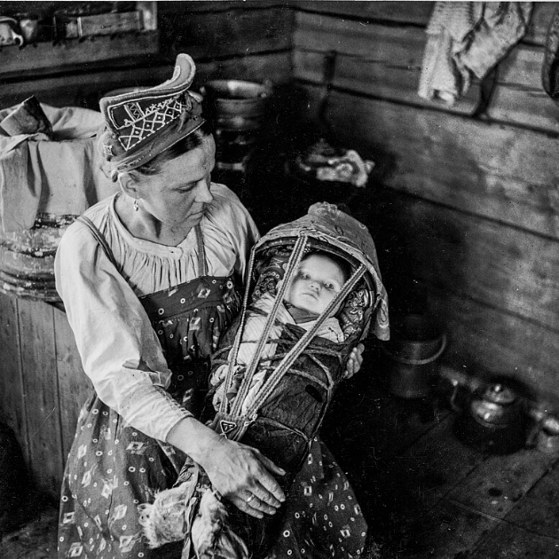 A black and white photo of a Sami mother in traditional clothing holding a baby swaddles in a cradleboard