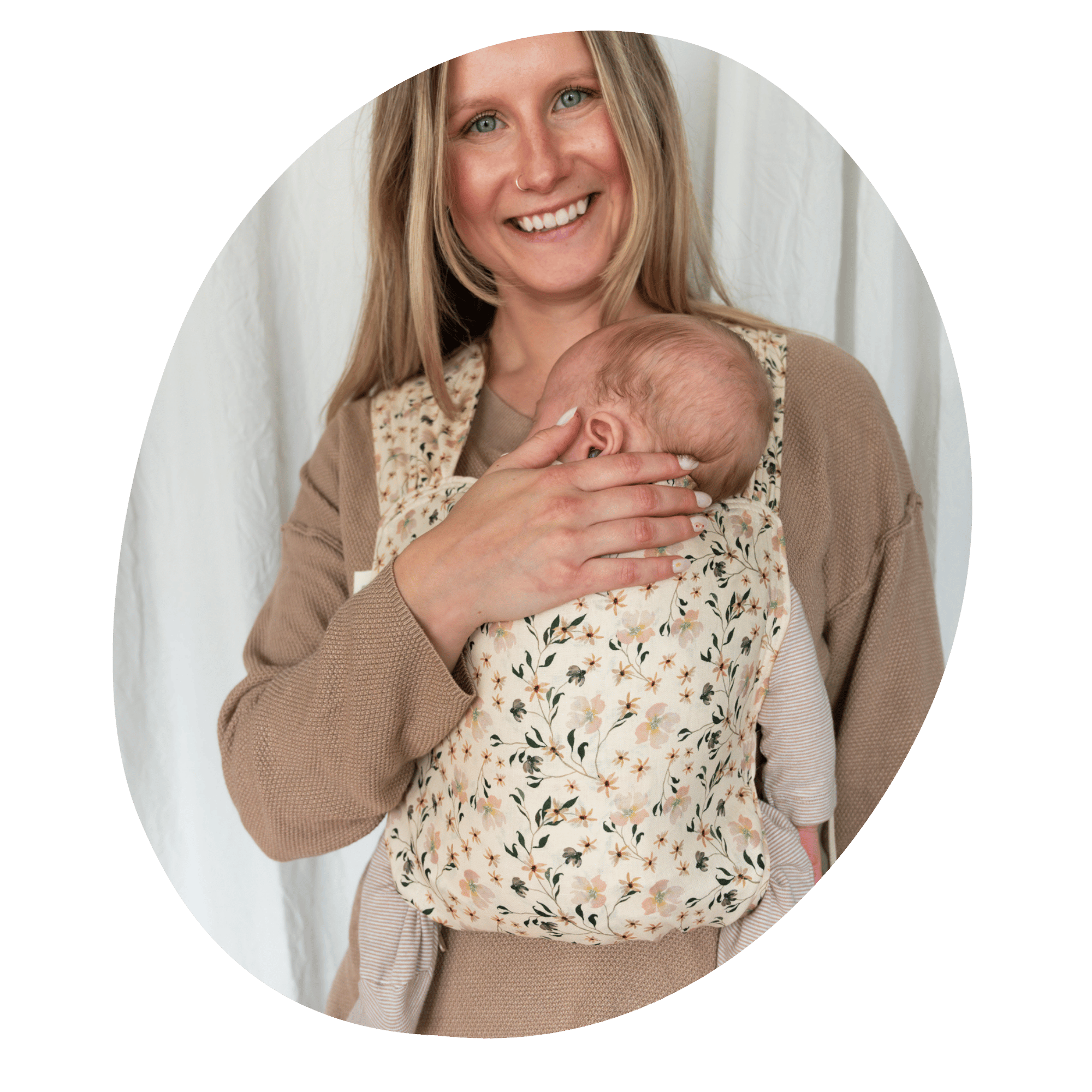 A white, straight size woman smiles at the camera while she embraces her newborn worn on her front in a Sprout
