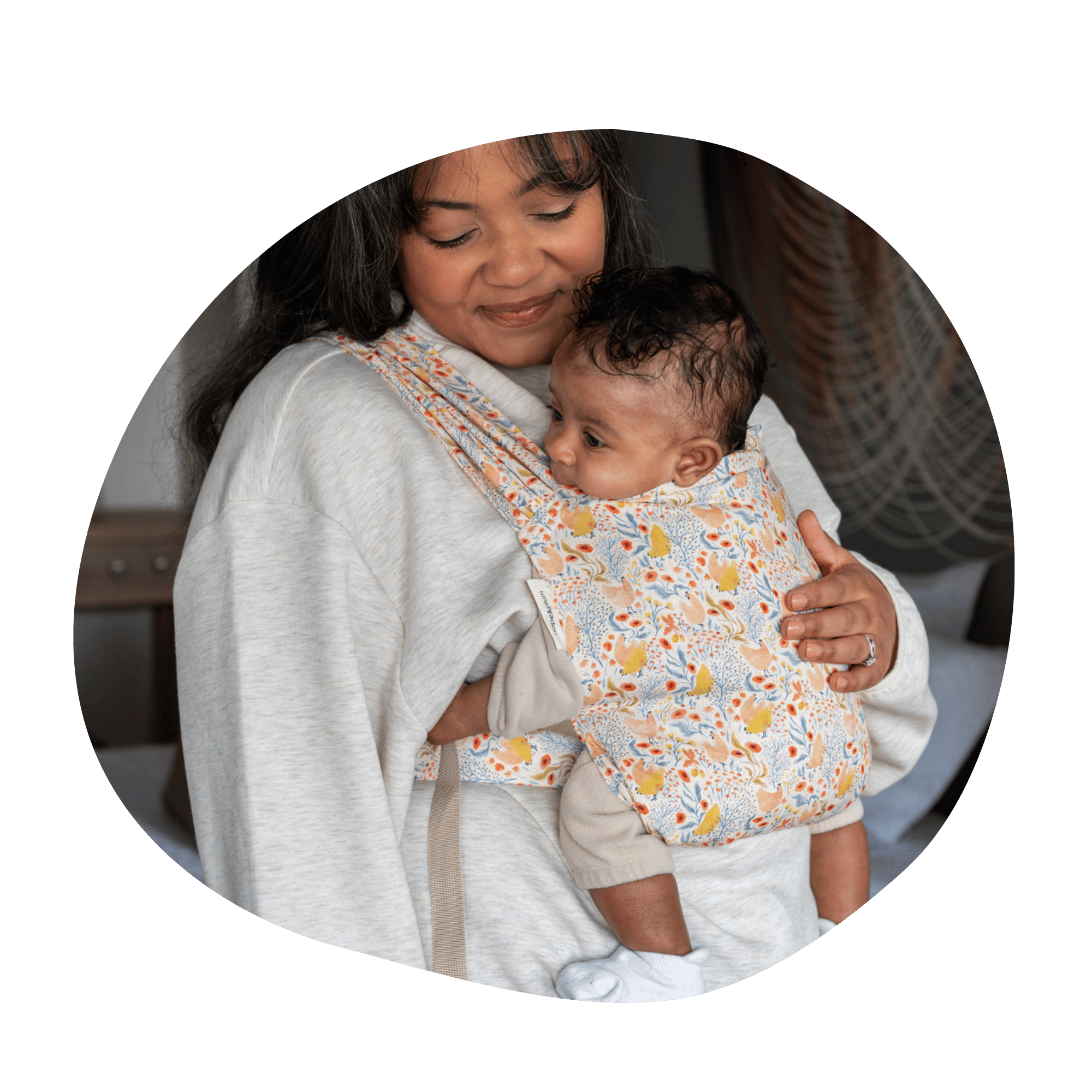 A plus size BIPOC woman smiles gently down at her infant worn on her front in a Sprout newborn carrier