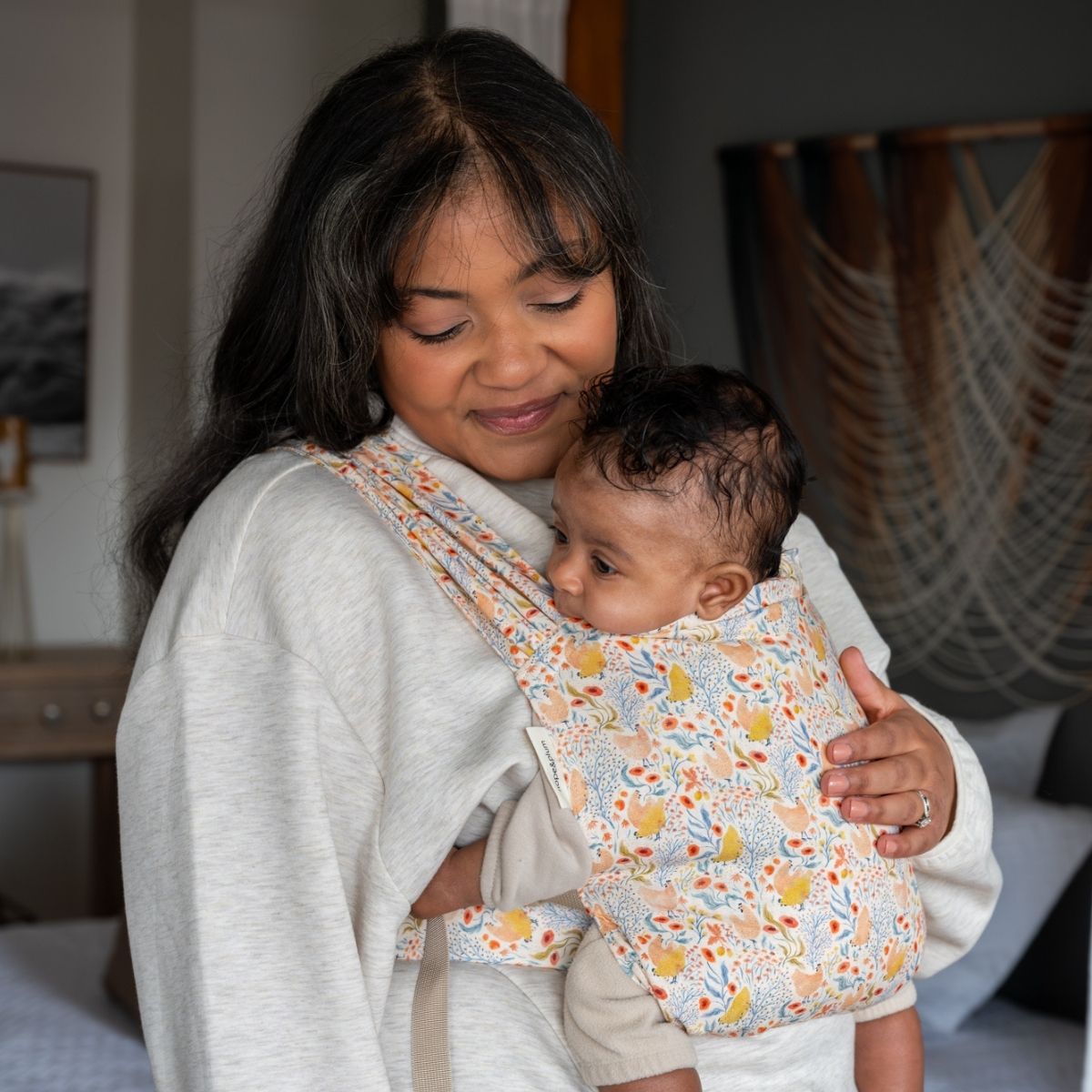 A plus size BIPOC woman smiles gently down at her infant worn on her front in a Sprout newborn carrier