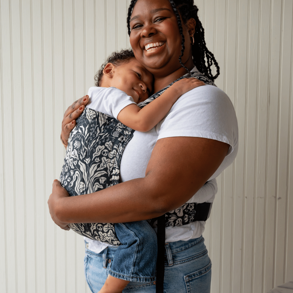 A mid size BIPOC woman smiles and wears her older baby in a hope&plum Lark on her front