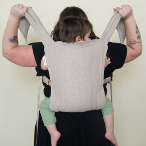 a plus size white woman holds both hope&plum Lark strap above her head to tighten her baby in a back carry