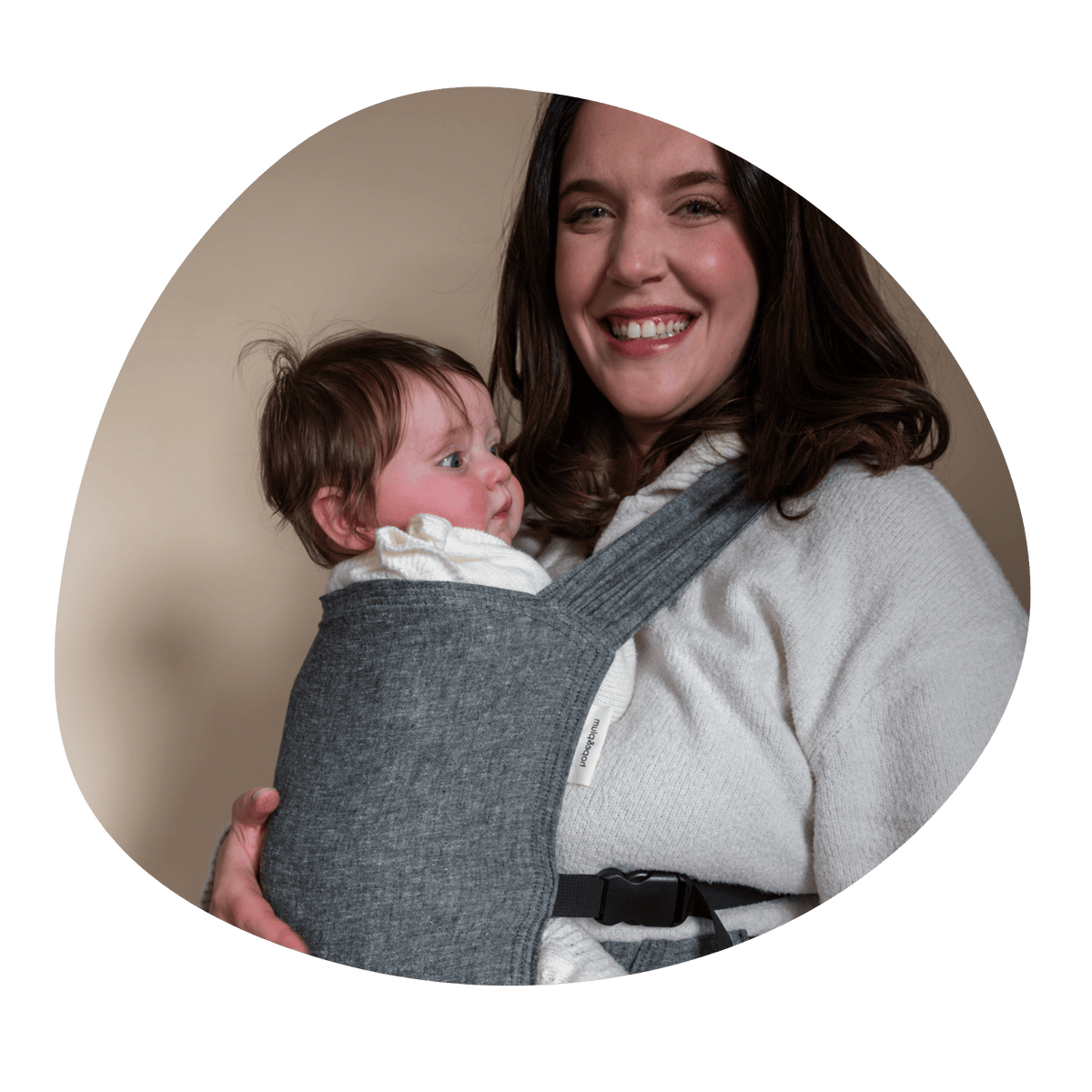 A white woman smiles at the camera while embracing her older infant worn on her front in a Sprout