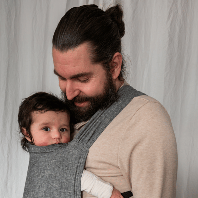 A white straight size bearded man smiles down at his infant worn on his front in a hope&plum Sprout Newborn Carrier
