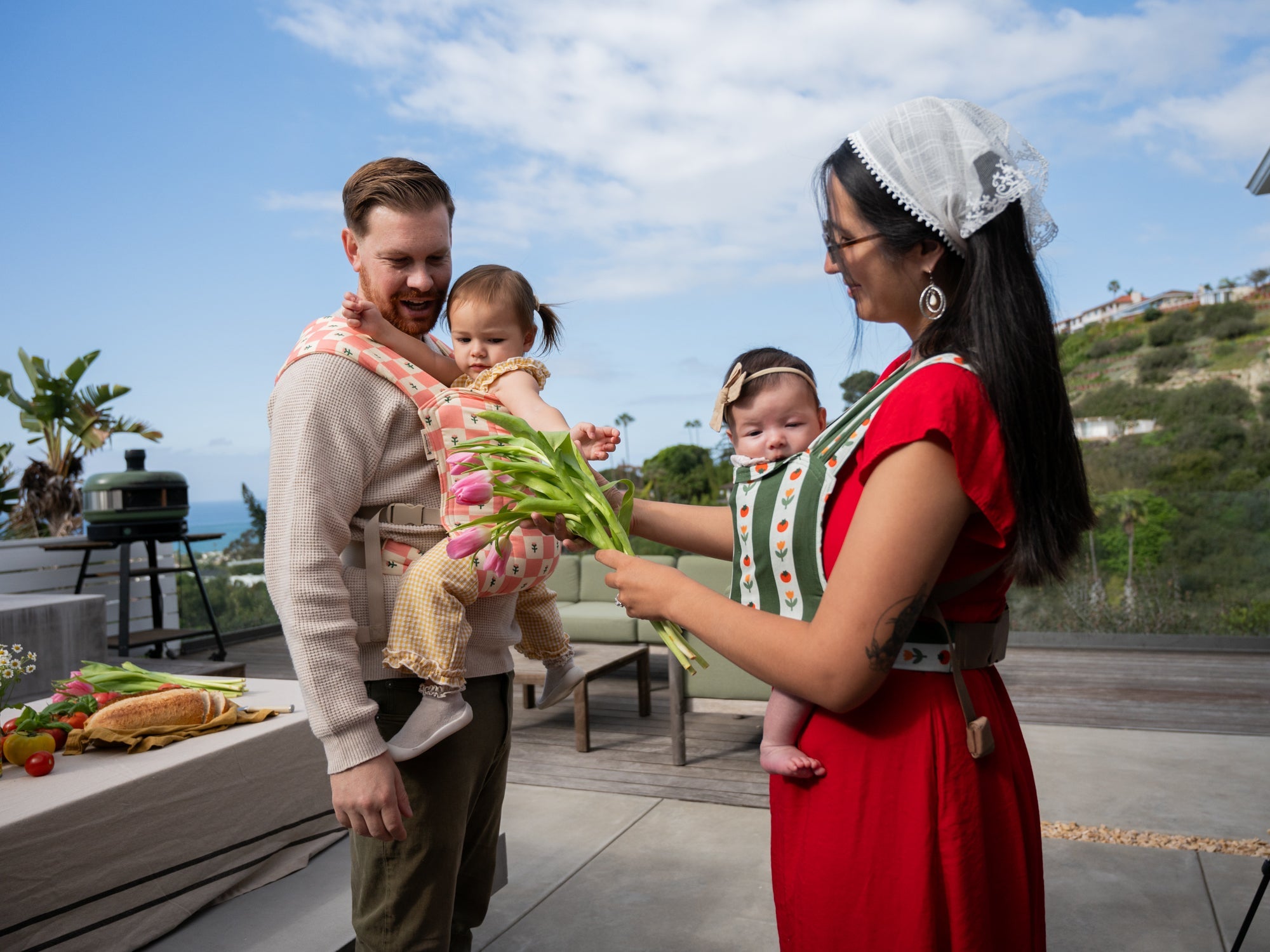 Three caregivers stand while carrying babies: one in a ring sling front carry, one in a back carry in a meh dai, and one in a stretchy wrap. The middle caregiver is the only one seen in full.
