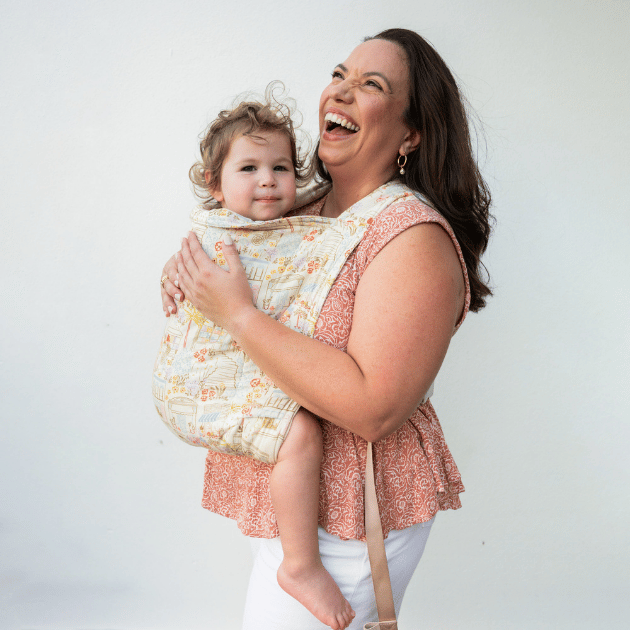A woman wears her baby in a kid Lark on her front. She looks upward and laughs while baby looks at the camera.