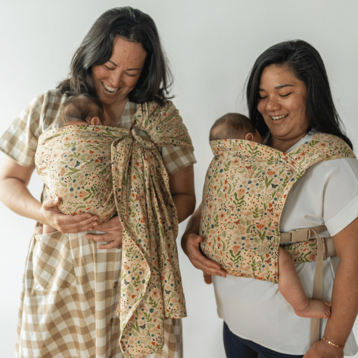 Two woman smiling down at their babies worn in floral pattern hope&plum carriers, one wrap and one Lark