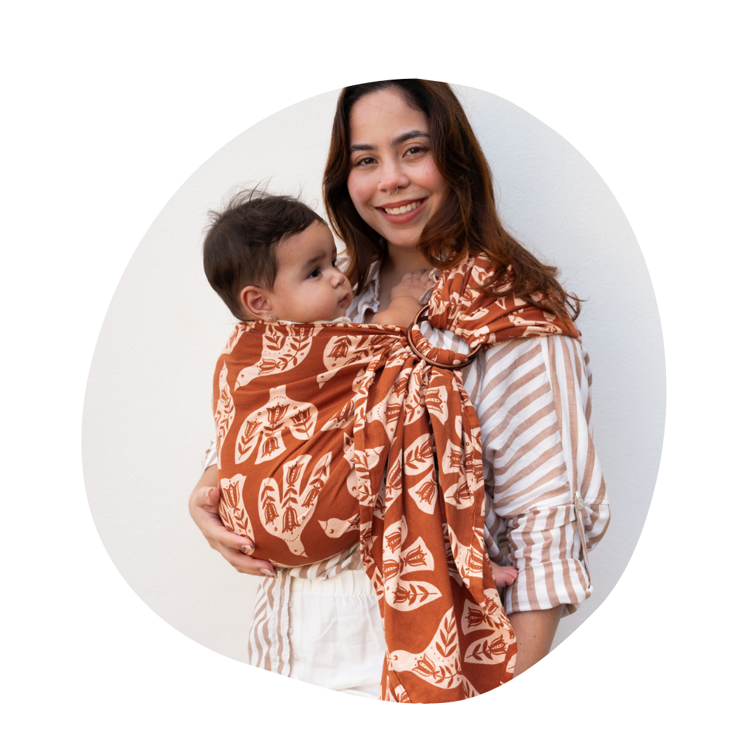 A woman stands while wearing a baby in a ring sling.