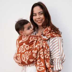 A woman stands while wearing a baby in a ring sling.