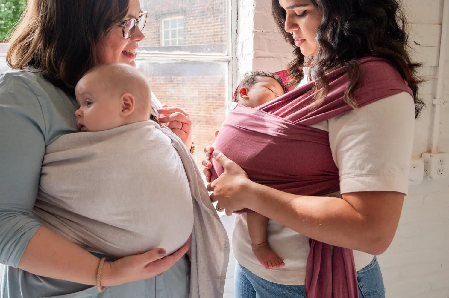 White mother wearing her baby in a gray ring sling talking to another mother wearing her newborn in a rose-colored baby wrap.