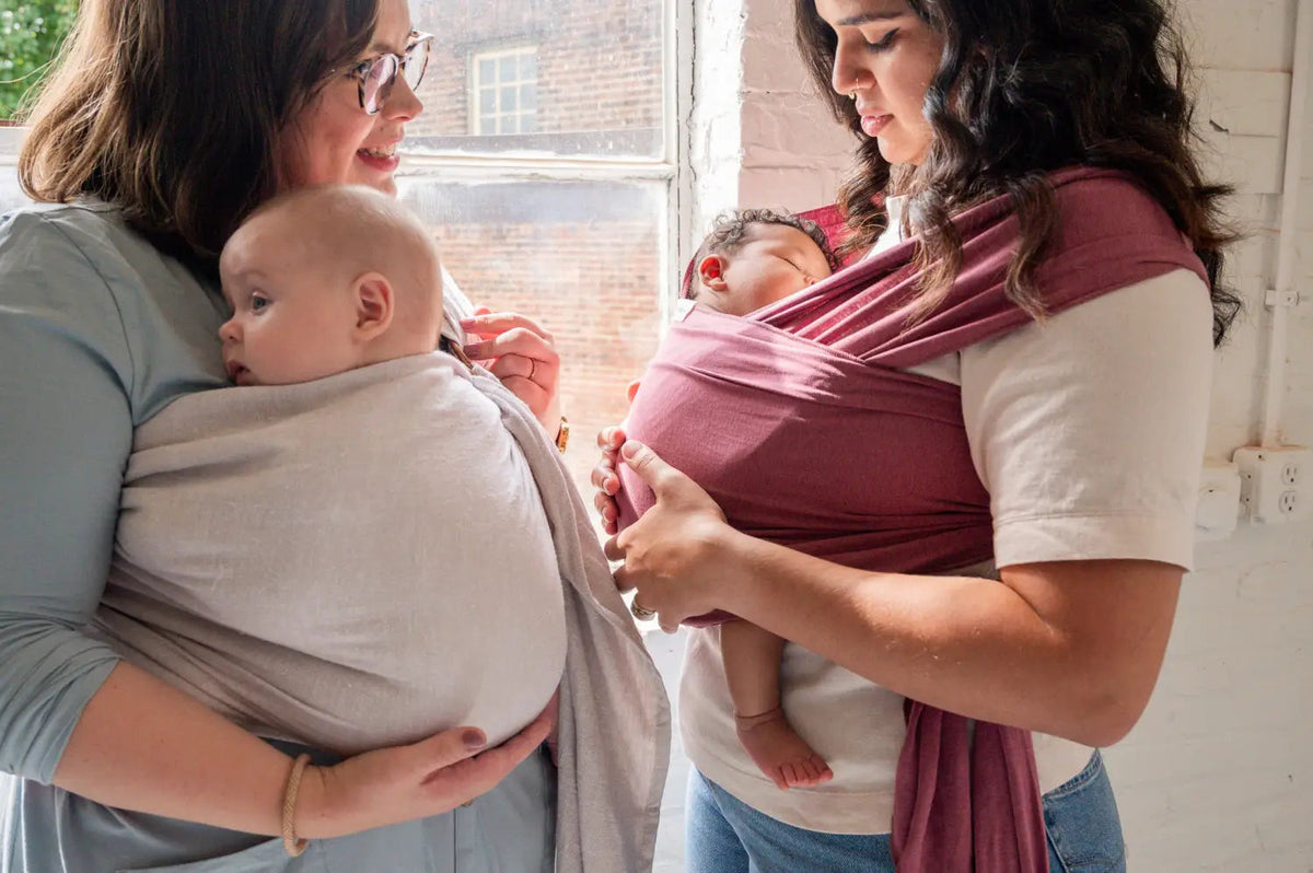 White mother wearing her baby in a gray ring sling talking to another mother wearing her newborn in a rose-colored baby wrap.