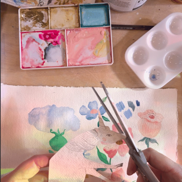 A close up on the design process for a print. A pair of white hands hold a paper unicorn and scissors above a watercolor painting of flowers