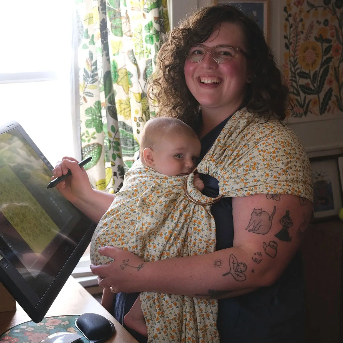 Phoebe Wahl, a plus-size white artist, smiles at the camera while working in her studio and wearing her baby on her front in a hope&plum ring sling
