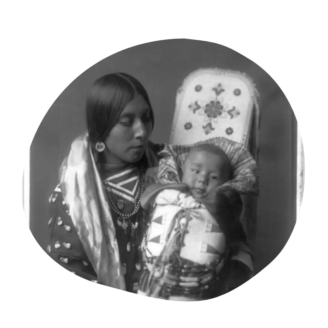 A black and white photo of a Native American woman dressed in traditional clothing sitting and holding up a baby in a cradleboard.