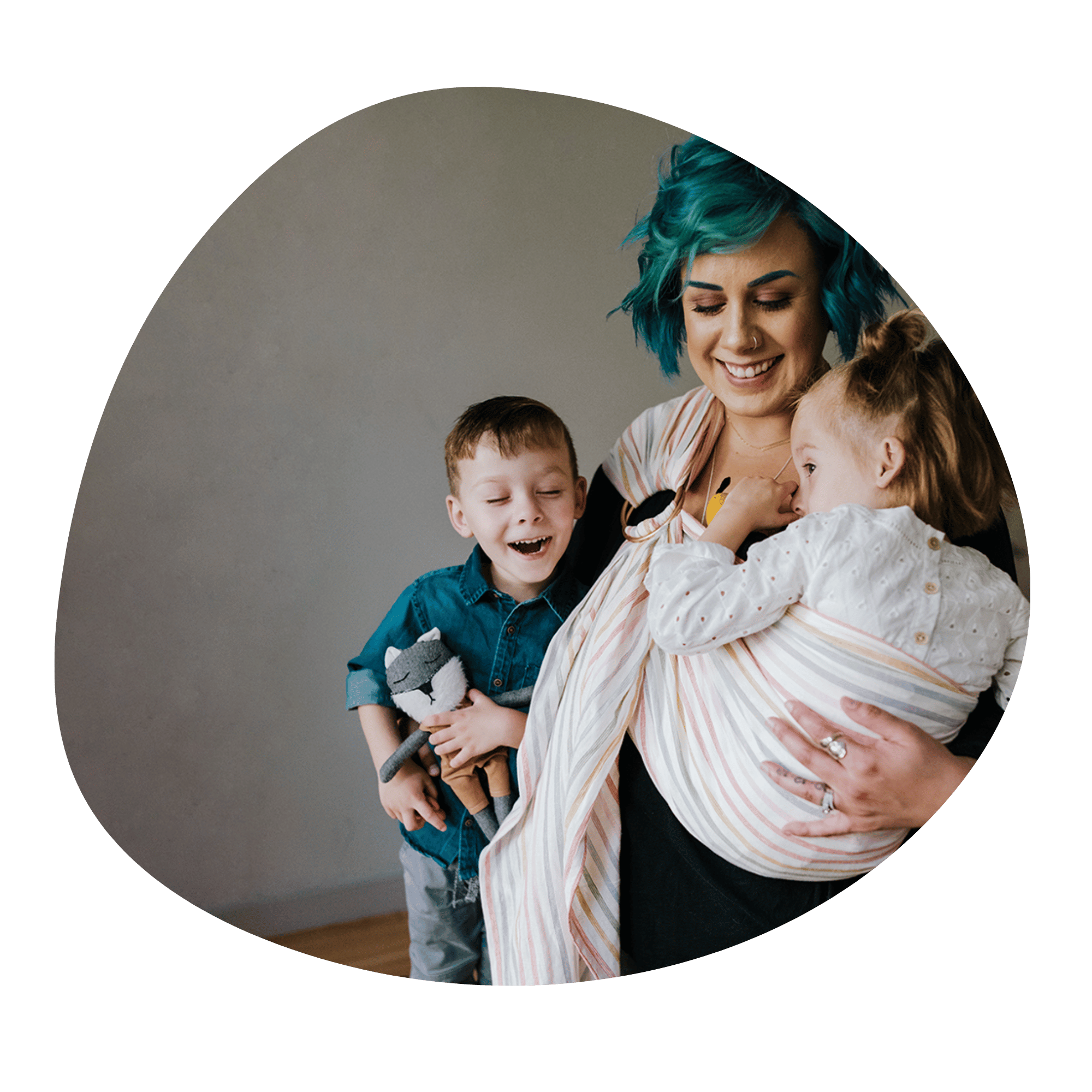 A light skinned woman with blue hair embraces a toddler she wears on her side in a ring sling. The toddler sucks her thumb. Standing close to them is a little kid smiling with his eyes closed.