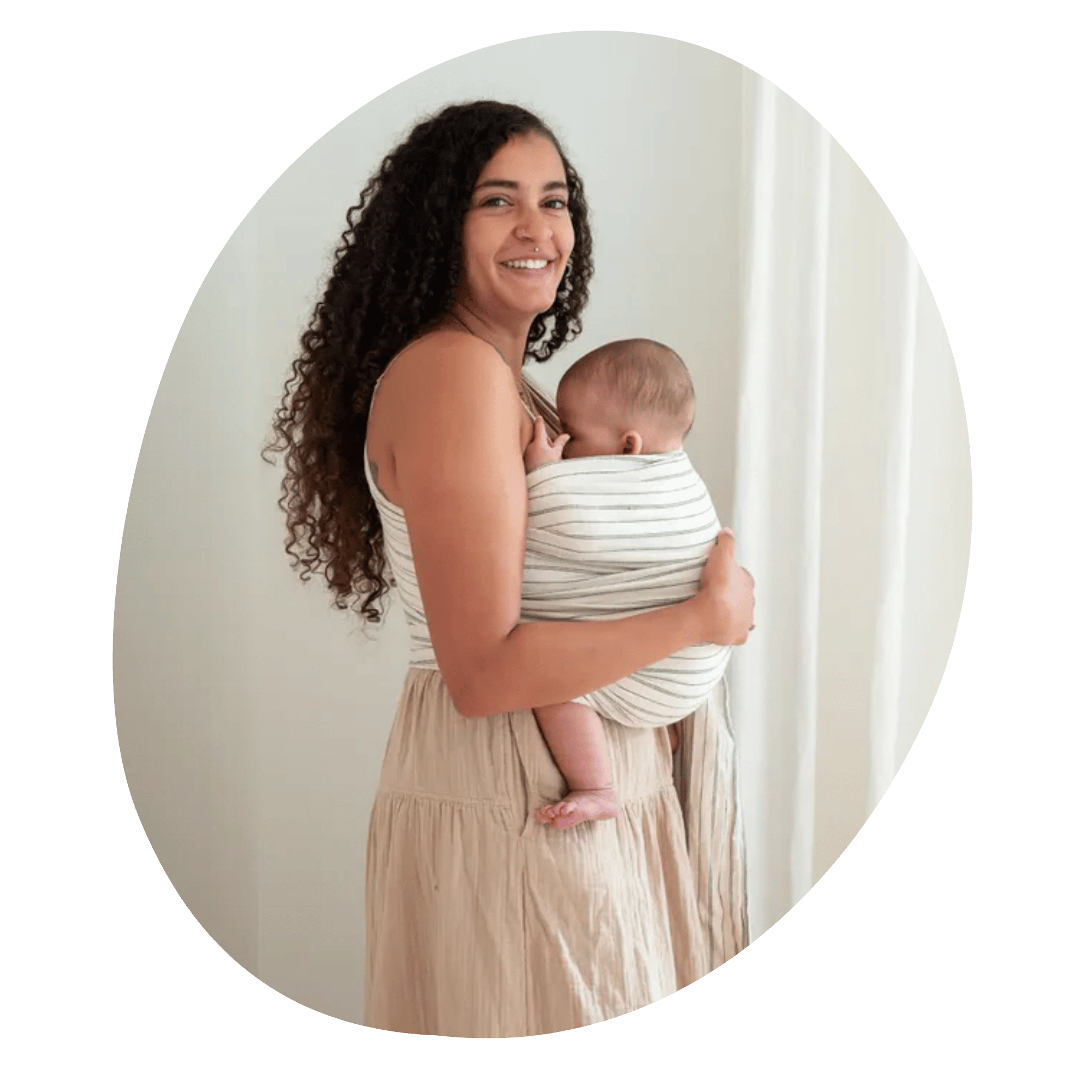 A brown skinned woman with brown curly hair embraces the brown skinned baby she wears on her front in a ring sling. She stands in profile, looks at the camera, and smiles.