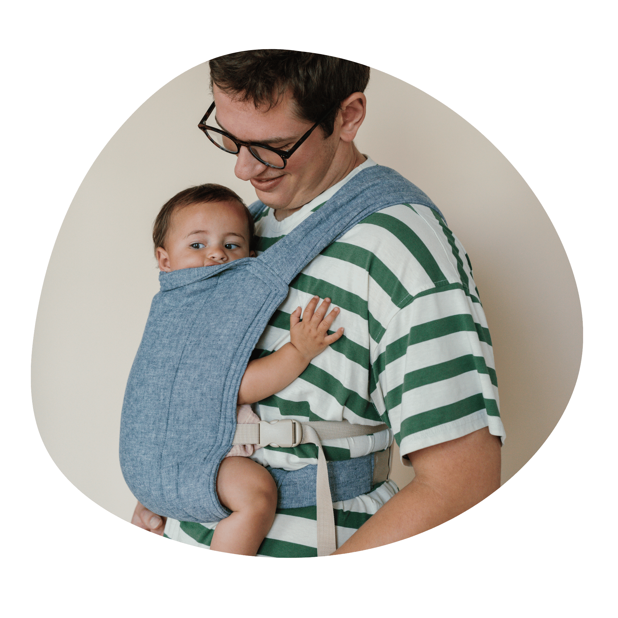 Man in a green striped shirt and glasses smiling and holding his baby in a blue chambray baby carrier