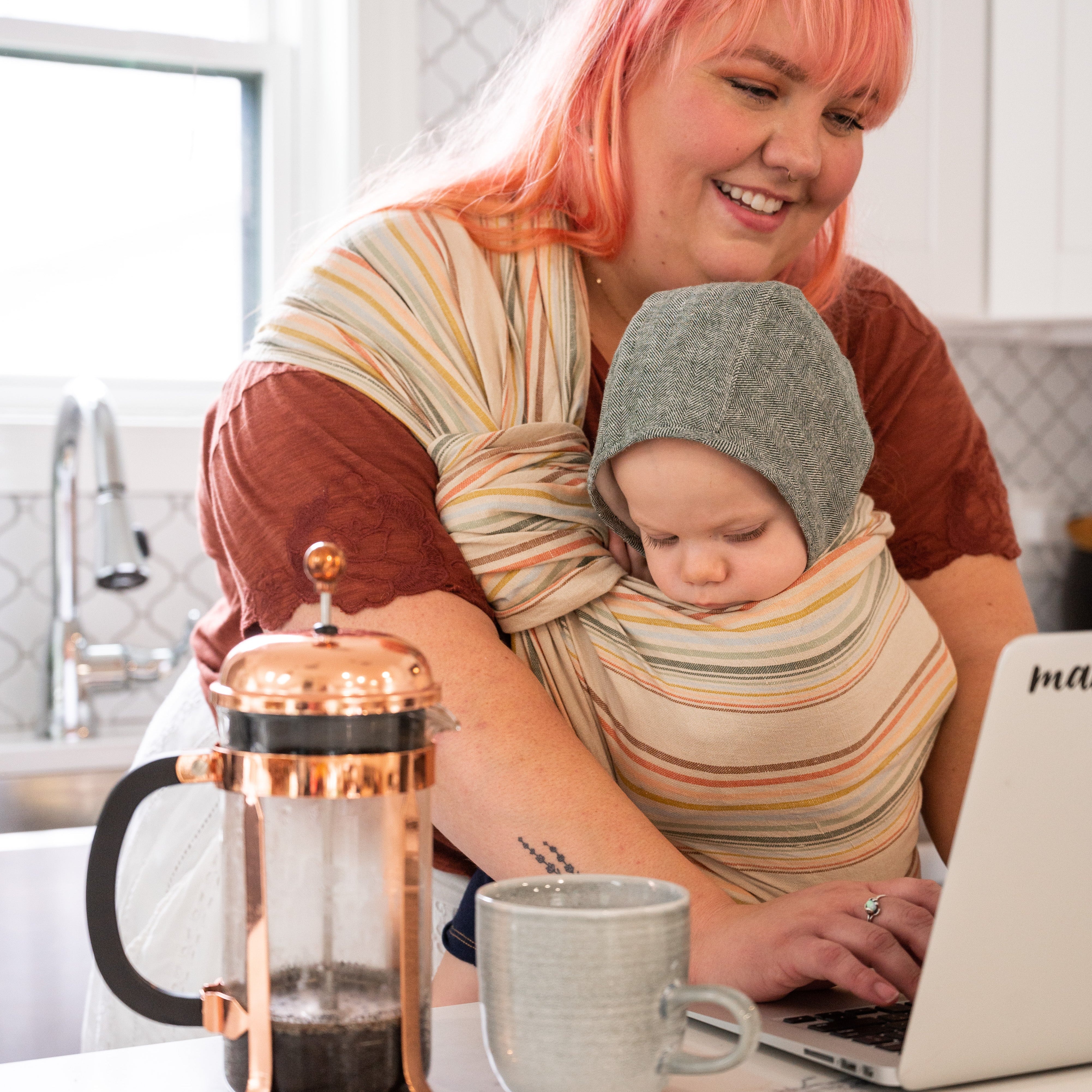 Woman with pink hair using a laptop while holding a baby in a sling, with a French press and mug on the table.