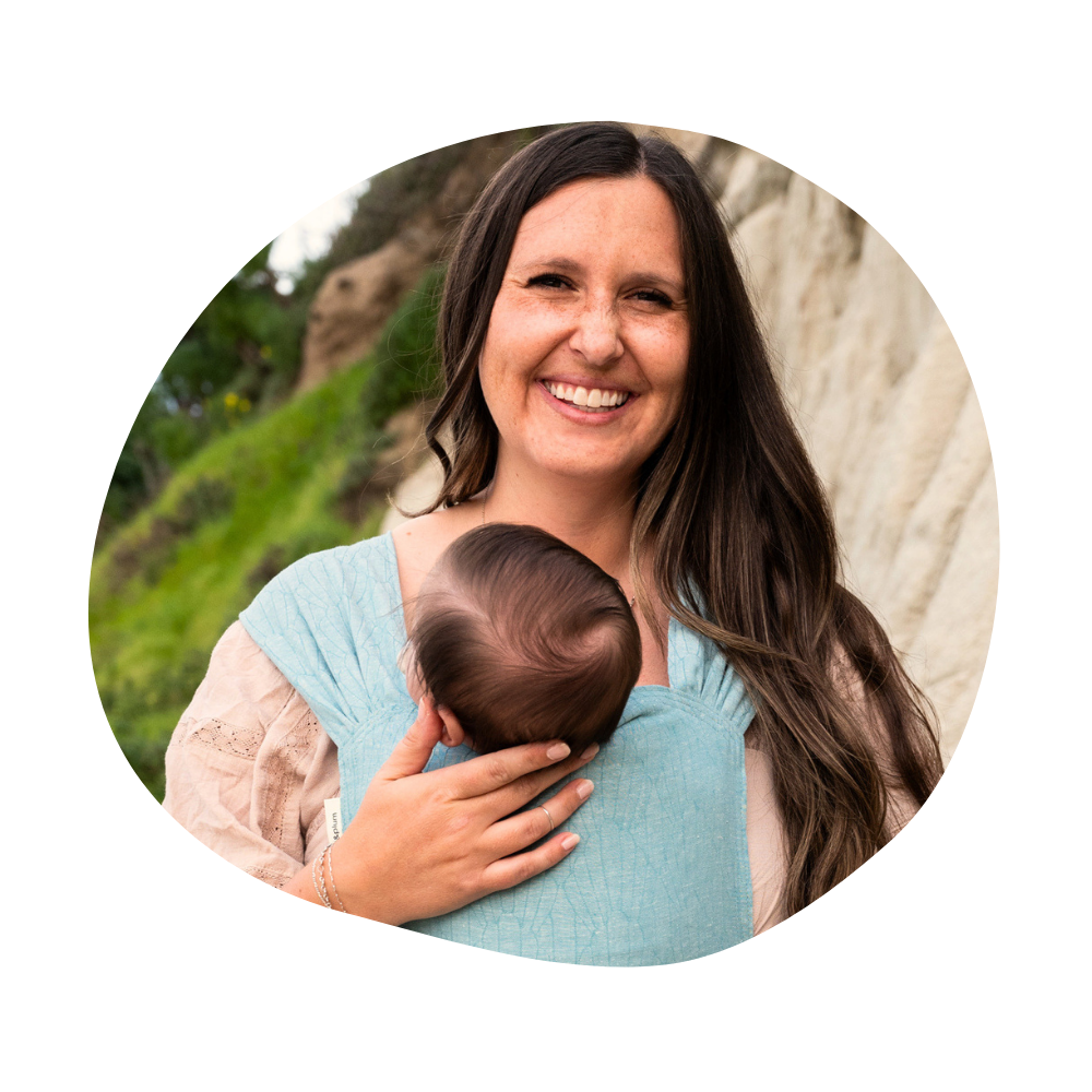 Kenzie, a smiling woman with long brown hair, carries a baby against her chest in a light-blue Maren Sprout carrier, standing outdoors beside a grassy cliff.