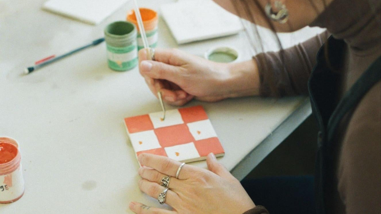 A close up photo of Toni Darling Frank painting a ceramic tile with a pink and white checkerboard pattern.