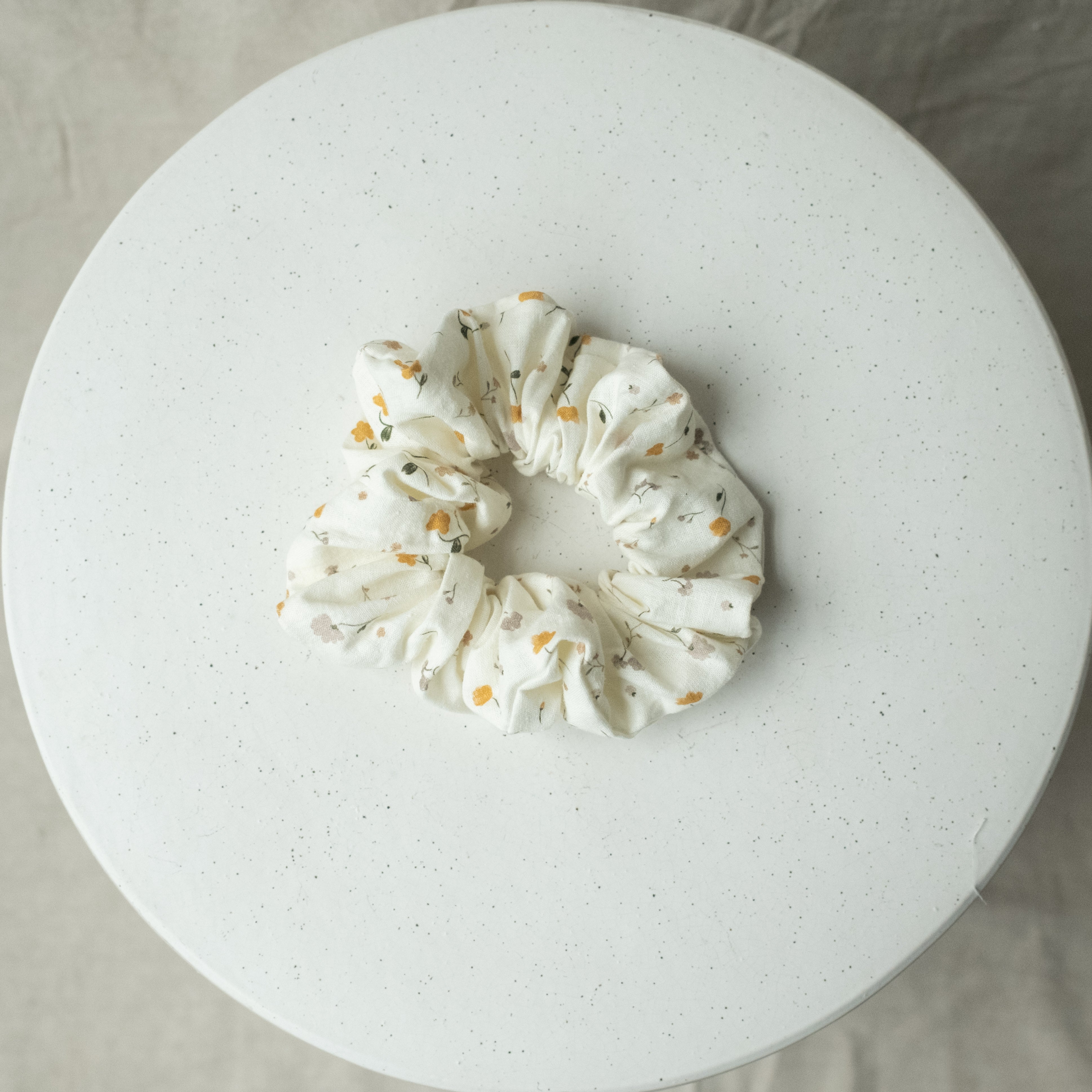 A single oversized scrunchie in white woven fabric printed with a delicate scattered blossom pattern in mustard yellow and blush pink with fine green stems.