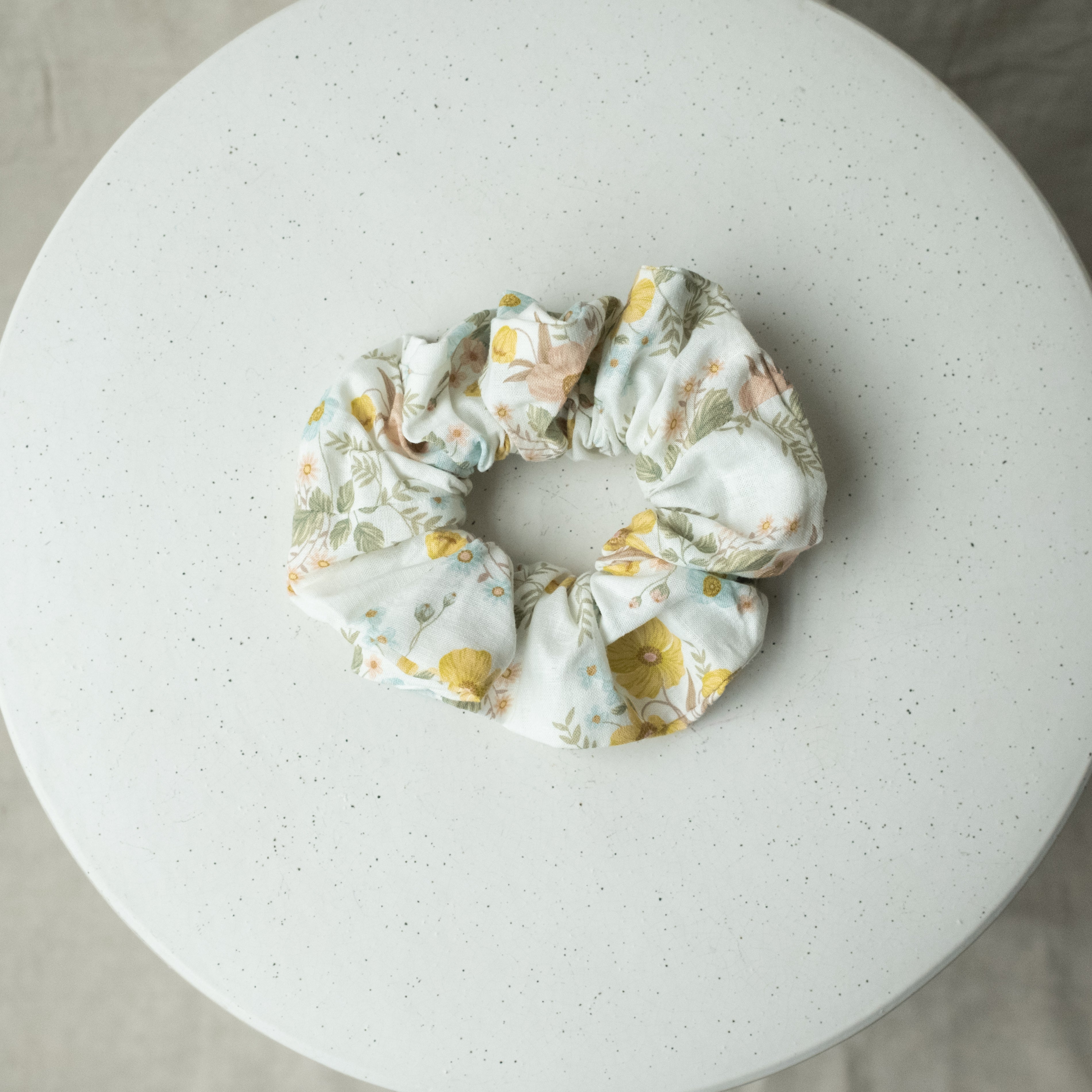 A single oversized scrunchie in white woven fabric printed with a soft botanical pattern of buttercups and foliage in mustard yellow, sage green, blush pink, and pale blue.