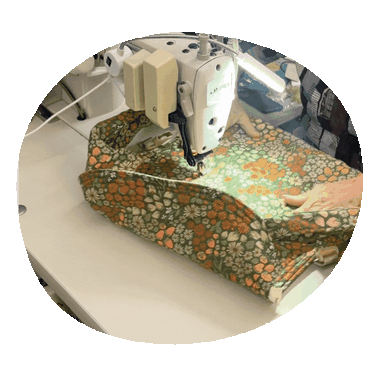 Animated GIF of hands guiding Fête fabric — a rich green woven print with orange, yellow, and white wildflowers — through an industrial sewing machine as a Lark carrier takes shape.
