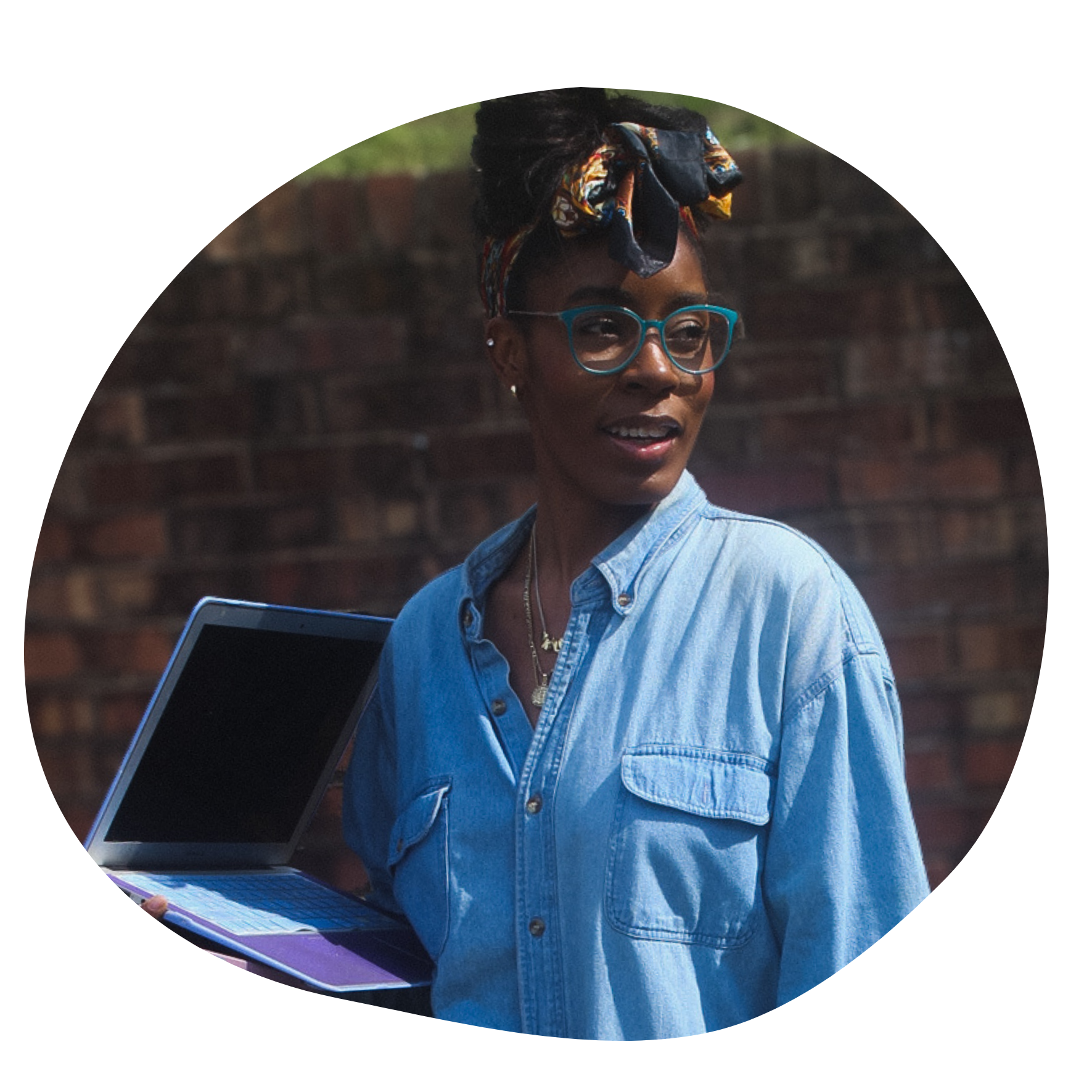A black woman wearing teal glassess with her long hair tied up in a silk scarf holds a laptop.