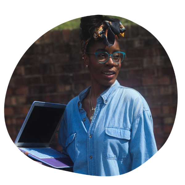 A black woman wearing teal glassess with her long hair tied up in a silk scarf holds a laptop.