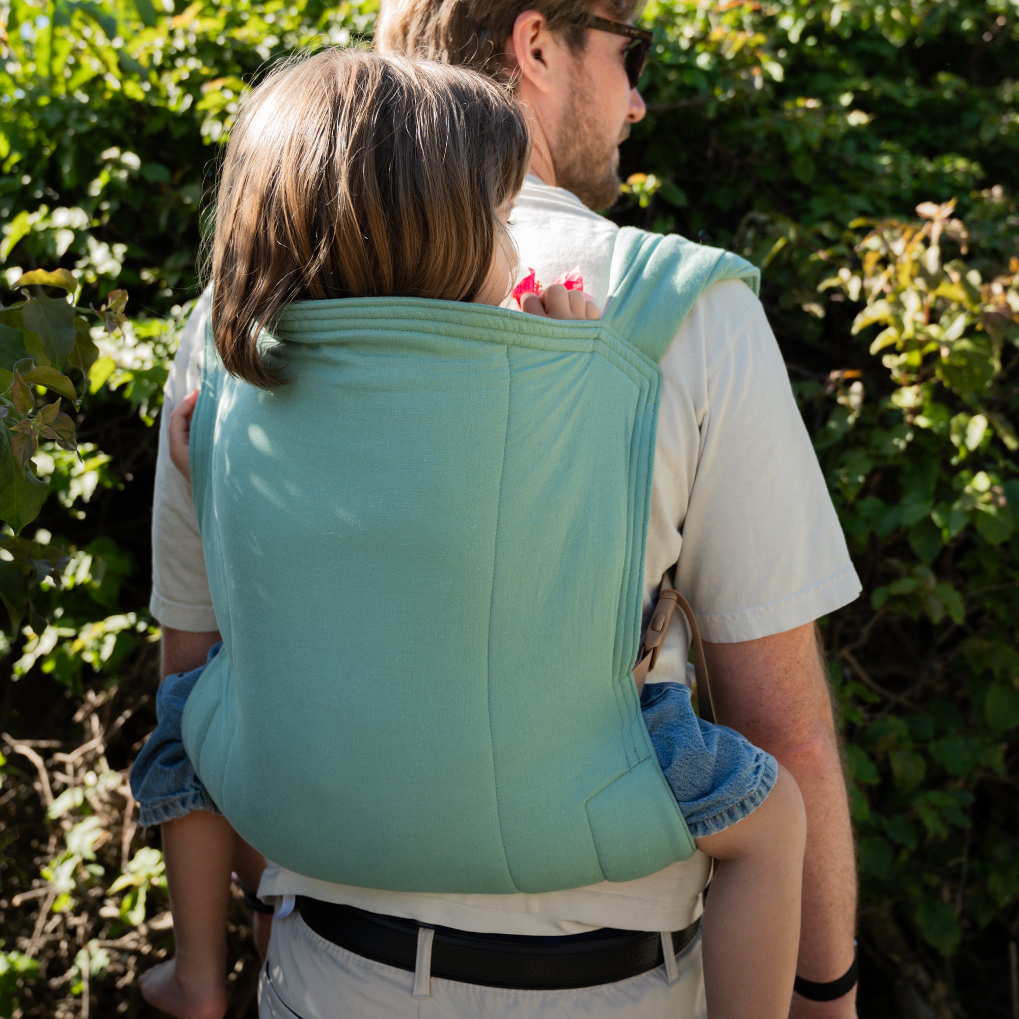 Person carrying a child in a green/blue baby carrier outdoors with greenery in the background