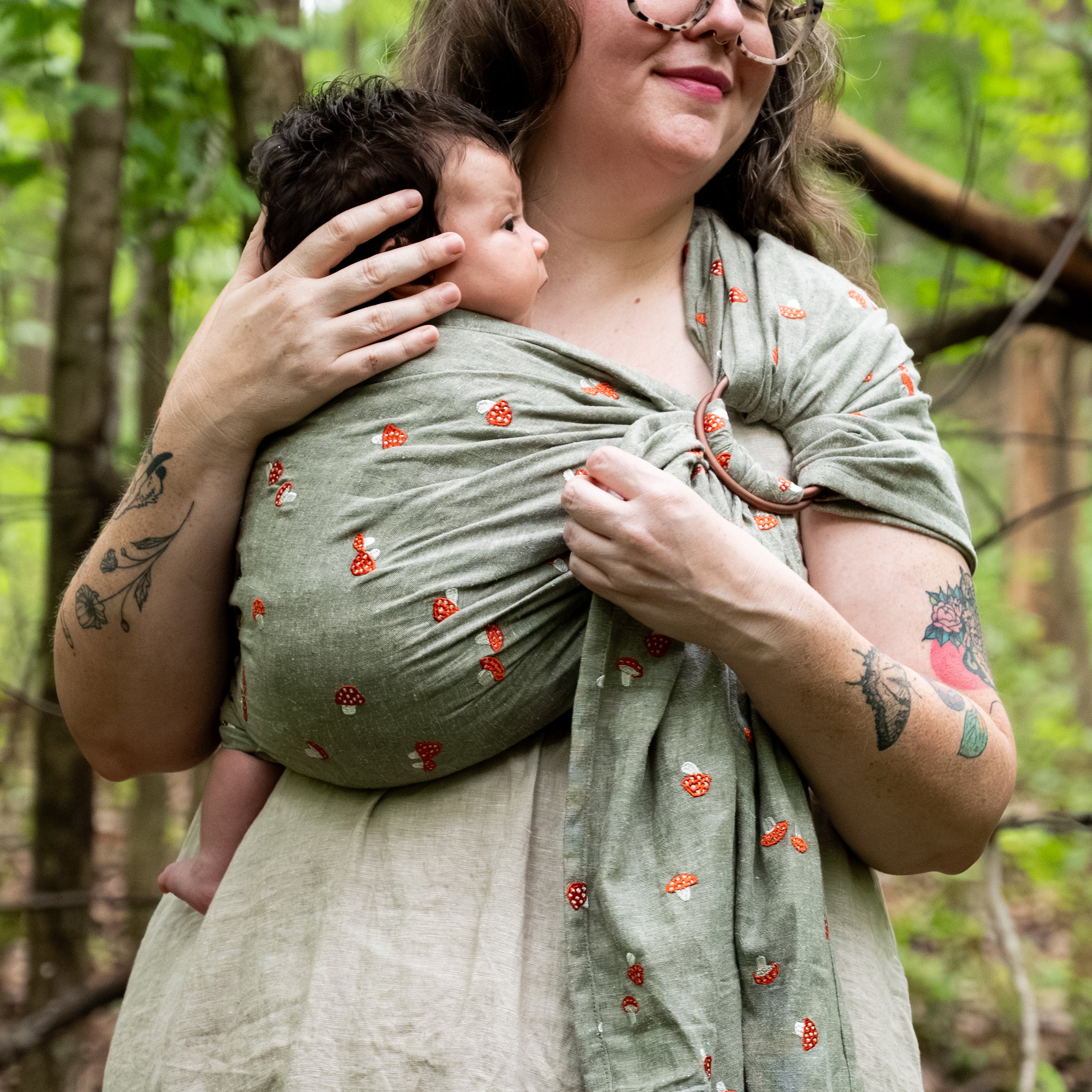 Woman in a wooded setting holding a newborn against her chest in the Amanita Ring Sling, our sage-green chambray fabric, Palm, embroidered with fly agaric red and white spotted mushrooms, with the shoulder ring visible.