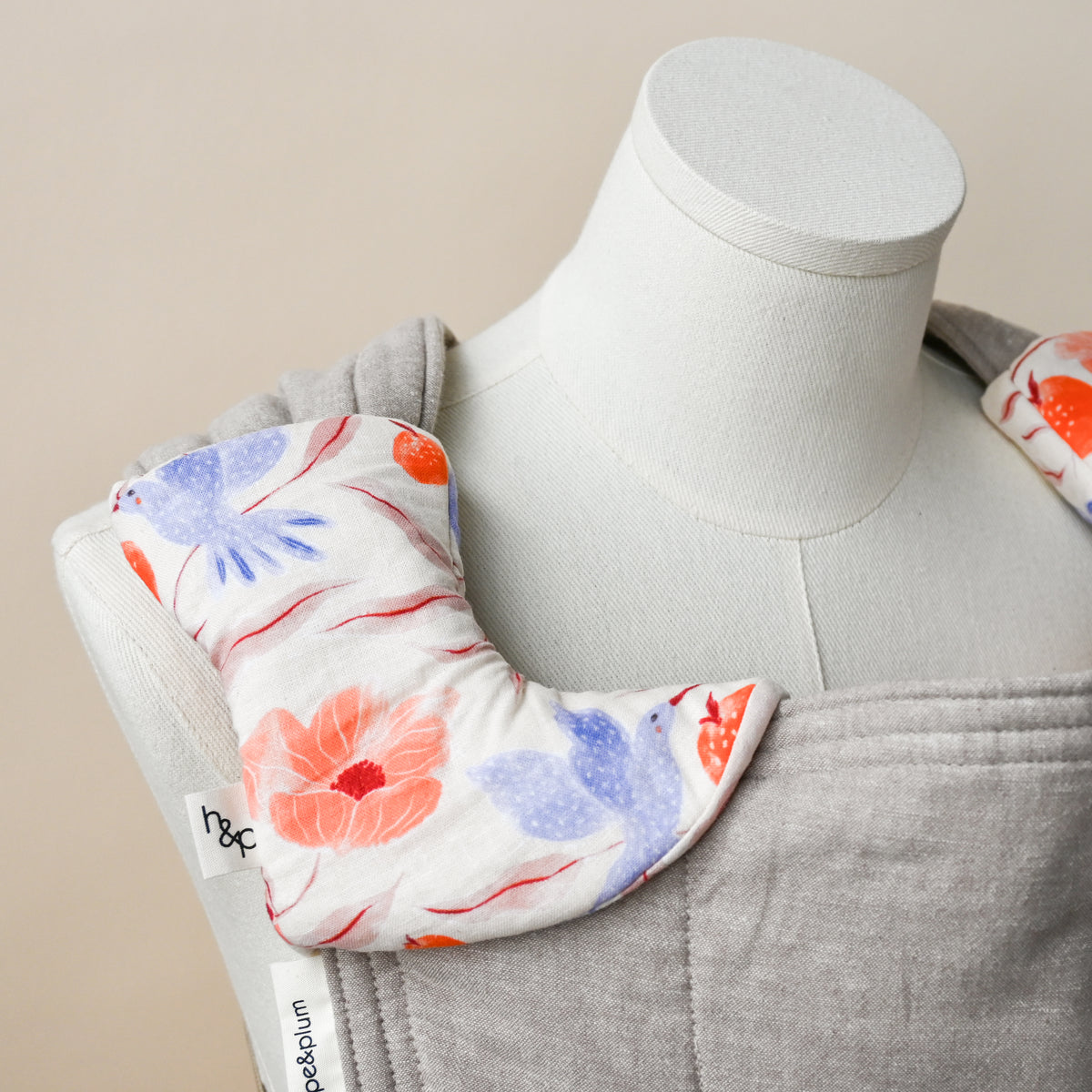 Drool Pads in Ambrosia, wrapped over a carrier shoulder strap on a dress form, cream fabric hand-painted with blue hummingbirds, orange pomegranates and coral poppies.