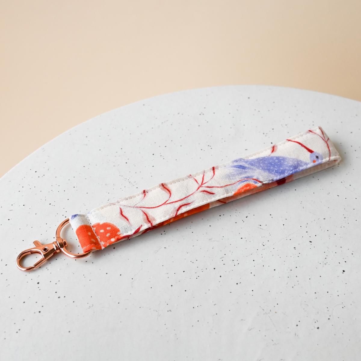 Key Fob in Ambrosia, a fabric wristlet strap with a rose gold clasp, cream fabric hand-painted with blue hummingbirds, orange pomegranates and coral poppies.