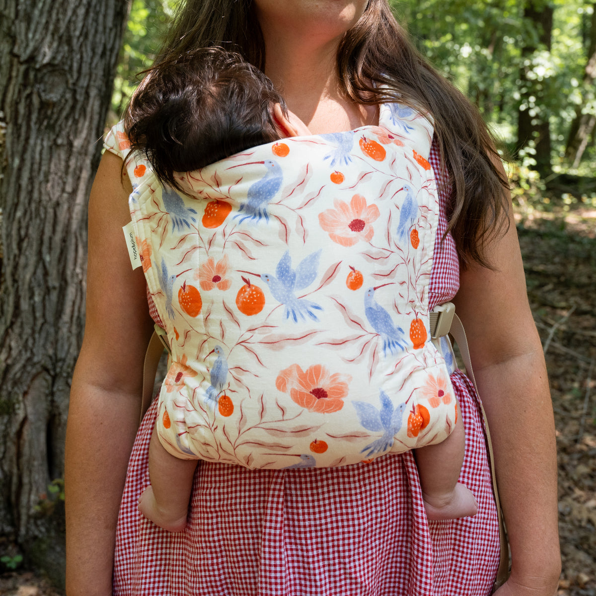 A baby rests in the Ambrosia Lark Baby Carrier worn in a front carry, the panel printed with blue hummingbirds, coral poppies and orange pomegranates on cream.