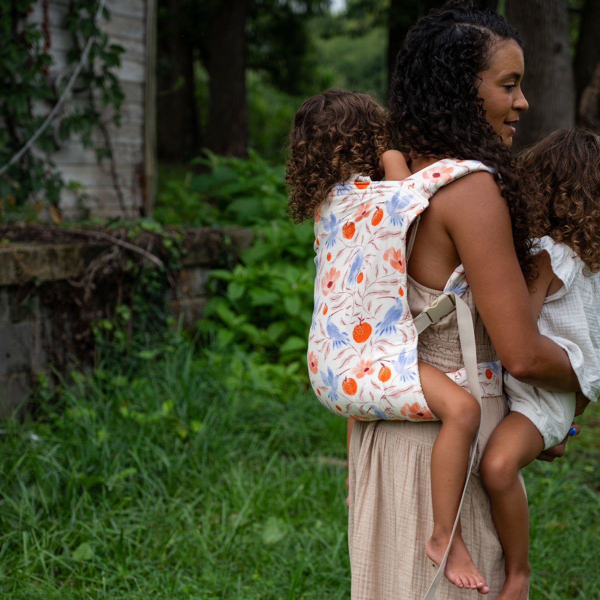 A child rides in a back carry in the Ambrosia Lark Kid Carrier while the wearer holds a second child on the hip, outdoors in long grass.
