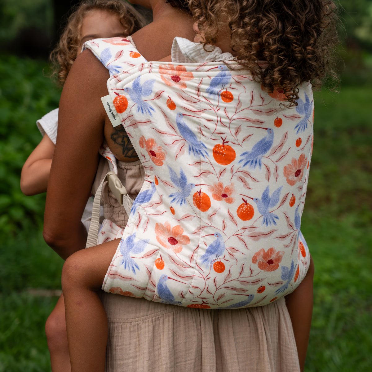 Back view of a child carried in the Ambrosia Lark Kid Carrier, the full panel of hummingbirds, poppies and pomegranates on cream across the wearer's back.