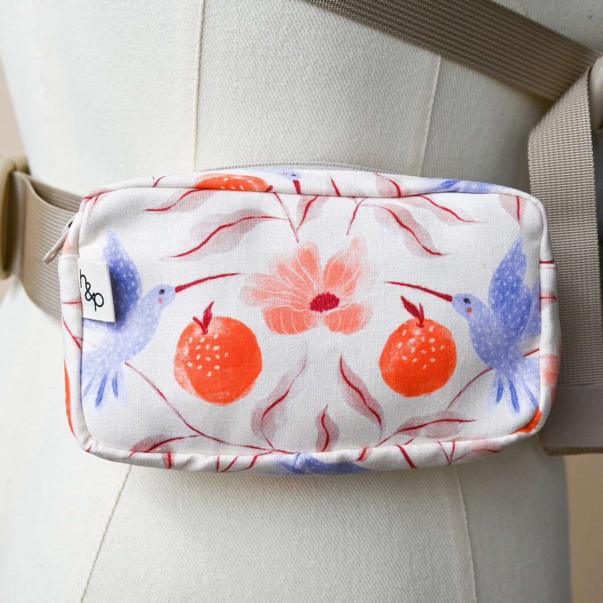Carrier Pocket in Ambrosia, a zip pouch on a carrier waistband shown on a dress form, cream fabric hand-painted with blue hummingbirds, orange pomegranates and coral poppies.