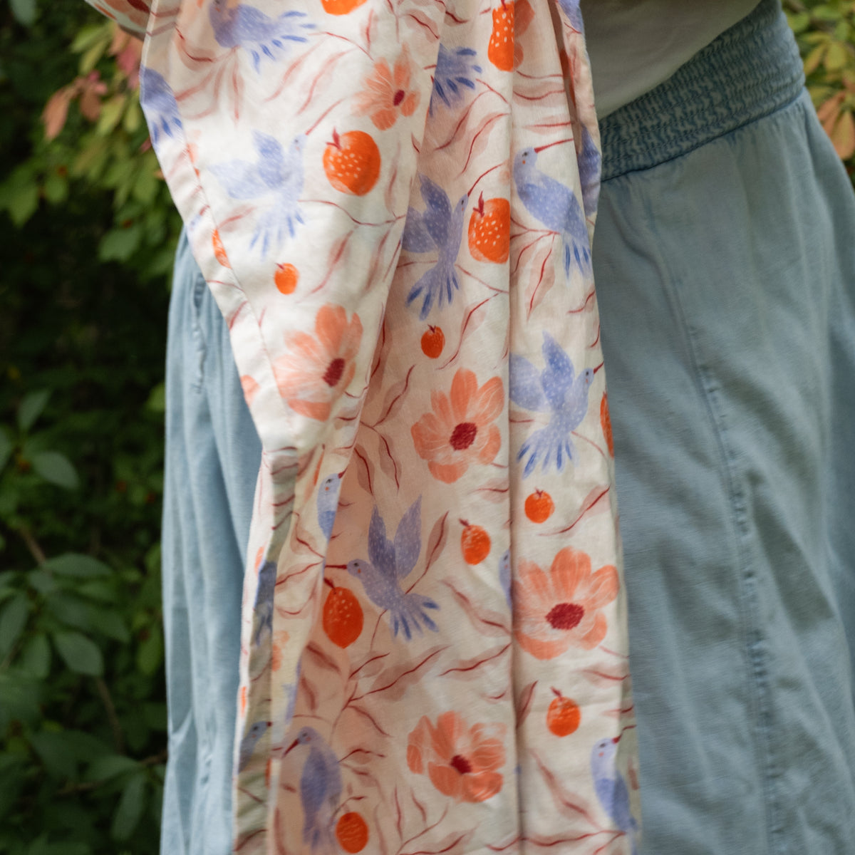 Close view of the Ambrosia fabric draping in folds, showing long-beaked blue hummingbirds among coral poppies, orange pomegranates and dusty rose leaves on cream.