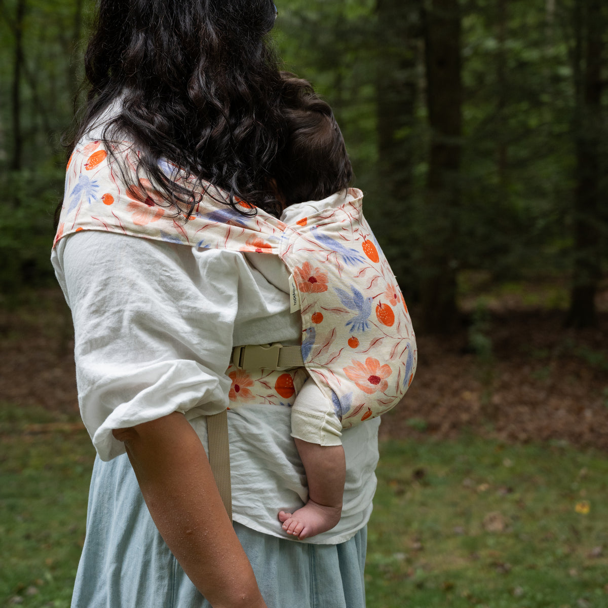 Side view of a baby carried in the Ambrosia Sprout Newborn Carrier, the printed panel drawn over the wearer's shoulder and across the back.