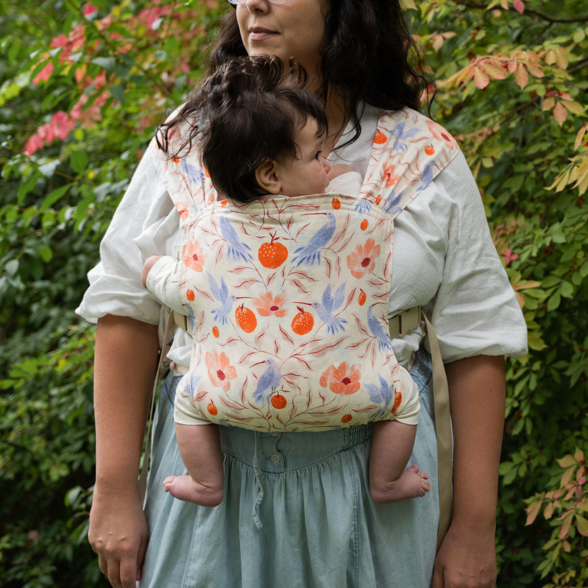 A wearer carries a baby in the Ambrosia Sprout Newborn Carrier, the full panel of blue hummingbirds, coral poppies and orange pomegranates on cream in view.